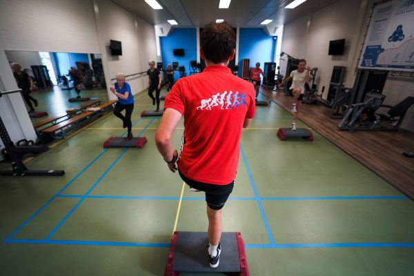 Een man geeft een steps-les in een sportstudio, waarbij deelnemers de bewegingen op de step volgen.