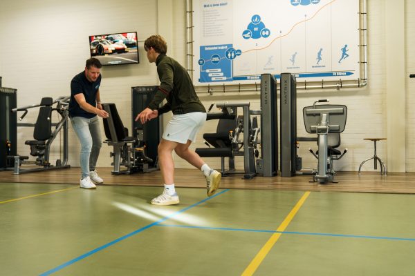 Fysiotherapeut geeft een jongen van 15 jaar instructies voor het uitvoeren van een stabiliteitsoefening gericht op landingstechniek in de oefenzaal.